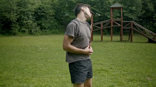 A young man is experiencing stomach discomfort or indigestion while standing in a park. He holds his stomach, stretches and breathes deeply, trying to relieve the pain or bloating he's feeling.
