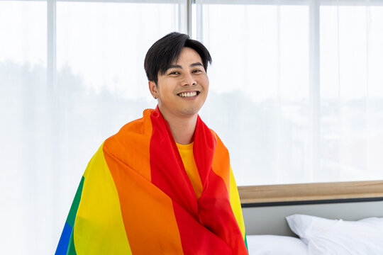 Smiling Asian LGBTQ gay man individual wrapped in a rainbow pride flag in a bright bedroom, looking at camera - Powered by Adobe