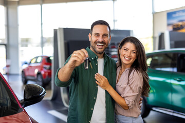 Visiting car dealership. Beautiful couple is holding a key of their new car, looking at camera and...