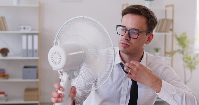 Slow motion video of upset young businessman sitting at desk in front of fan working in office and suffering from summer heat. Man employee using electric fan during heatwave in office. 4k video.