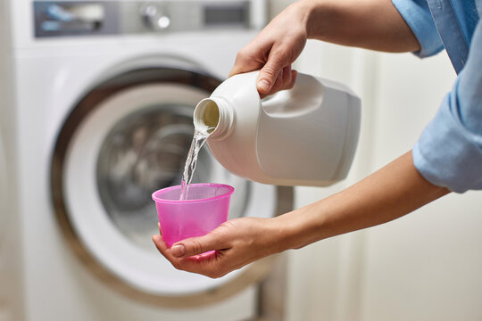Woman pours liquid transparent laundry detergent or conditioner into plastic pink cap against blurred washing machine.