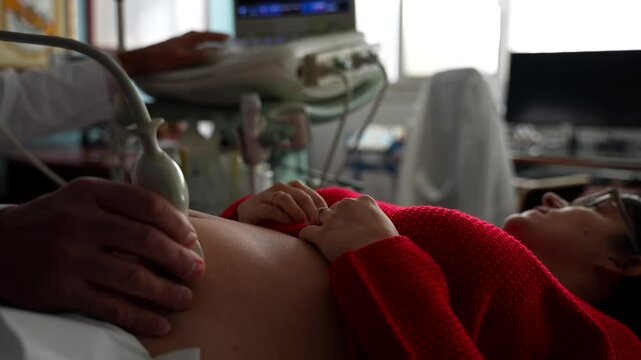 Pregnant woman lying on a stretcher looking at ultrasound screen while gynecologist scans her belly