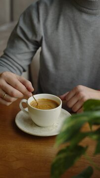 Close-up of male person sits comfortably at small table in cafe stirring coffee. Hands of unrecognized man in grey sweater mixing sugar in coffee cup.