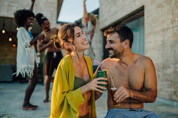 Happy couple drinking cocktails at a pool party with friends dancing in the background