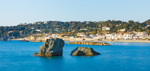 Panoramic view of Forio on Ischia Island, Italy, with rock formation Scoglio Dell'Impiccato in foreground. Clear blue sea, sandy beach with restaurants, sun lounger rentals and water sports facilities