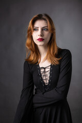 Fototapeta premium Red-haired gothic girl in a black dress on a gloomy background in the studio.