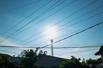 Tangled overhead power lines against a clear blue sky with a telecom tower in the background. Urban infrastructure, chaotic electrical wiring, and connectivity concept.