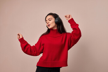 Happy excited woman wearing red sweater and headphones enjoying listening music and dancing on pink...