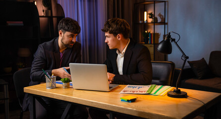 Young businessman in the office working late at evening, sitting at desk using laptop, talking with male assistant, checking reading paper documents.