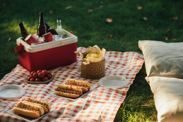 Picnic with hot dogs and cold drinks in cooler bag on red tablecloth in a park.