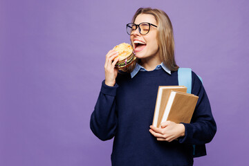 Young happy hungry girl student wear blue casual clothes glasses backpack bag hold books eating biting burger isolated on plain pastel light purple background. High school university college concept.