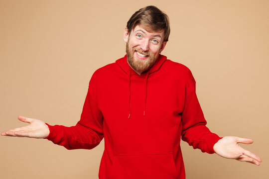 Fototapeta Young Caucasian man he wear red hoodie casual clothes shrugging shoulders looking puzzled, have no idea spread hands isolated on pastel plain light beige background studio portrait. Lifestyle concept.