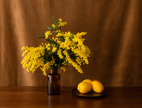 Bouquet de mimosa jaune dans vase en verre ambr&eacute;, avec trois citrons sur un plateau bronze pos&eacute; sur une table brune, vue de face avec un &eacute;clairage doux sur fond brun dor&eacute;