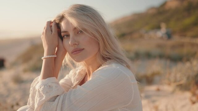 A beautiful young blonde woman with blue eyes and a white dress sits on a beach at golden hour and looks directly into the camera.