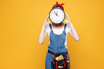 Young employee laborer handyman woman wearing overalls belt hold cover face with clock isolated on...