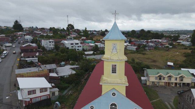 Aerial view of Colorful wooden church in Chonchi, Chiloe Island, a UNESCO World Heritage site, standing out against the backdrop of traditional houses and cloudy sky.circle dolly