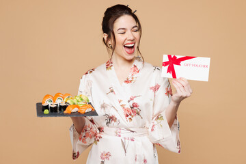 Young shocked happy woman wear kimono clothes hold eat raw fresh sushi roll served on black plate gift certificate coupon voucher card isolated on plain beige background studio. Japanese food concept.