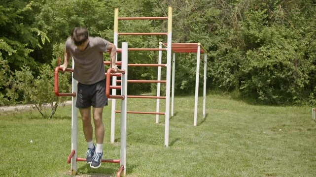 A young man performs dips on parallel bars in an outdoor park setting. He completes his set and walks away.