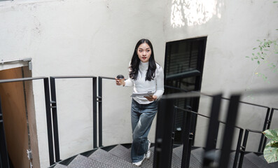 Young Asian woman walking up spiral staircase with coffee and documents, balancing work and daily routine.
