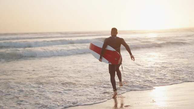 A surfer in a wetsuit runs into the ocean waves at sunset with his surfboard. He is ready to surf.