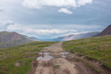 Dramatic landscape with puddle of water on dirt road on grassy flowering hill in wide alpine valley. Sunlit long mountain range far away under gray cloudy sky. Changeable weather in high mountains.
