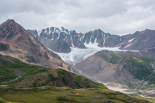 Dramatic landscape with orange tent in alpine valley with view to big glacier tongue and large sharp snow-capped mountain range under gray cloudy sky. Green hills and rocks against ice and sheer crags