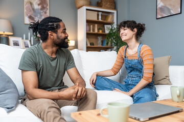 Smiling millennial couple sit on sofa in living room involved in interesting pleasant conversation at home, happy young man and woman talk chat relaxing on couch, enjoy romantic weekend or date