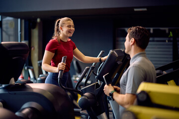 Happy athlete talking to her coach while working out on cross trainer in gym.
