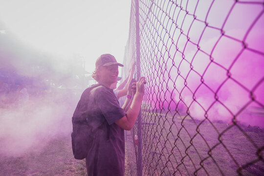 Teenagers watching vehicle burnout competition through chain mesh fence with smoke haze from burning