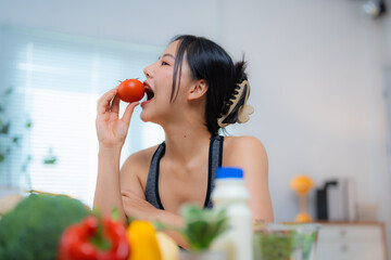 Young woman biting a tomato while promoting healthy eating and active lifestyle, surrounded by fresh vegetables and healthy foods in her kitchen