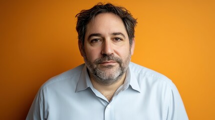 Obraz premium A middle-aged man with a beard and mustache wearing a light blue shirt stands confidently. His serious expression contrasts with the vibrant orange backdrop, creating an engaging atmosphere
