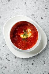 A close-up of a white bowl filled with vibrant red borscht soup, garnished with herbs and small dumplings, served on a minimalist plate against a textured gray background.