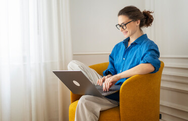 Relaxed happy smiling young woman freelancer in glasses and blue shirt relaxing and using laptop...