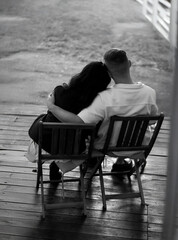 
A black and white photo of a couple sitting on chairs, with their backs to the camera, embracing. The girl rests her head on the guy&rsquo;s shoulder. An atmospheric photo.