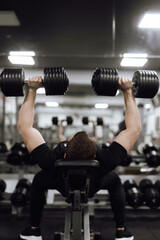 A man with an athletic physique lies on an inclined bench, lifting heavy dumbbells, showcasing...
