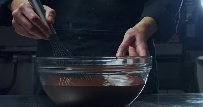Professional Chef mixing dark or milk Chocolate in a transparent bowl. Dessert maker prepare ingredient for cake on kitchen table. Chocolate drip and fall from steel whisk.