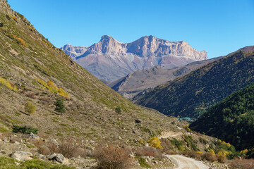Rugged mountain landscape with steep slopes and rocky terrain, featuring winding dirt road and patches of green vegetation under clear blue sky. Kabardino-Balkaria, Chegemsky district