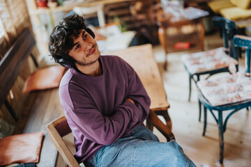 A young man sits comfortably, wearing headphones, in a cozy cafe. He appears relaxed and content,...