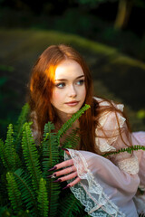 Beautiful red head girl in pink  vintage dress  standing near green plants and fern. Art work of...