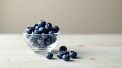 A glass bowl brimming with plump, juicy blueberries sits on a light wood surface, a few berries scattered nearby, evoking a sense of freshness and natural sweetness.