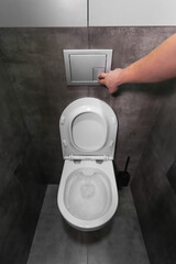 Man is pushing the flush button on a modern, wall-mounted toilet in a bathroom with contemporary grey tiles, highlighting hygiene and sanitation practices. Vertical photo