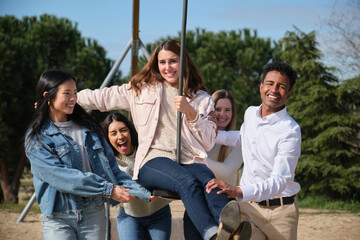 Five multi ethnic young adults enjoying time together at a park, pushing their friend on a swing