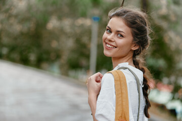 Cheerful young woman with a natural smile wearing casual white clothing and holding a tote bag, walking in a green nature setting, perfect for spring and summer concepts