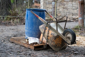 Orange wheelbarrow and blue barrel on wooden pallet in front of drystone wall
