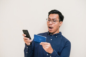 A Southeast Asian man is surprised to see his savings book he was holding and the other hand holding handphone on an isolated cream background, bank and finance concept