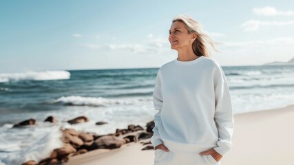 Fototapeta premium White sweatshirt mockup worn by a mature woman standing on a beach. The soft natural light and serene ocean backdrop. Ideal for showcasing apparel designs