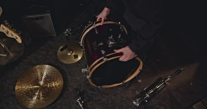 A drummer prepares his bass drum for a performance in a dimly lit room. He wipes the drum head and assembles the kick drum pedal.