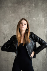 Fototapeta premium Confident woman in a black bodysuit and leather jacket, posing against a gray concrete wall, wearing black boots