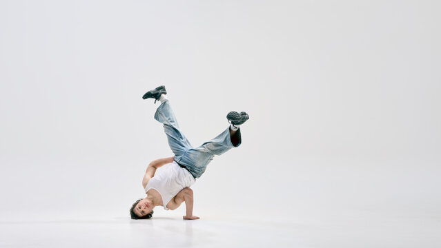 Dynamic photo of talented breakdancer balancing upside down on one hand with legs bent in air against white background. Concept of sport, art, music, strength, active lifestyle, rhythm, movement