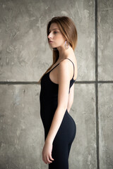 Fototapeta premium Confident woman in a black bodysuit and leather jacket, posing against a gray concrete wall, wearing black boots